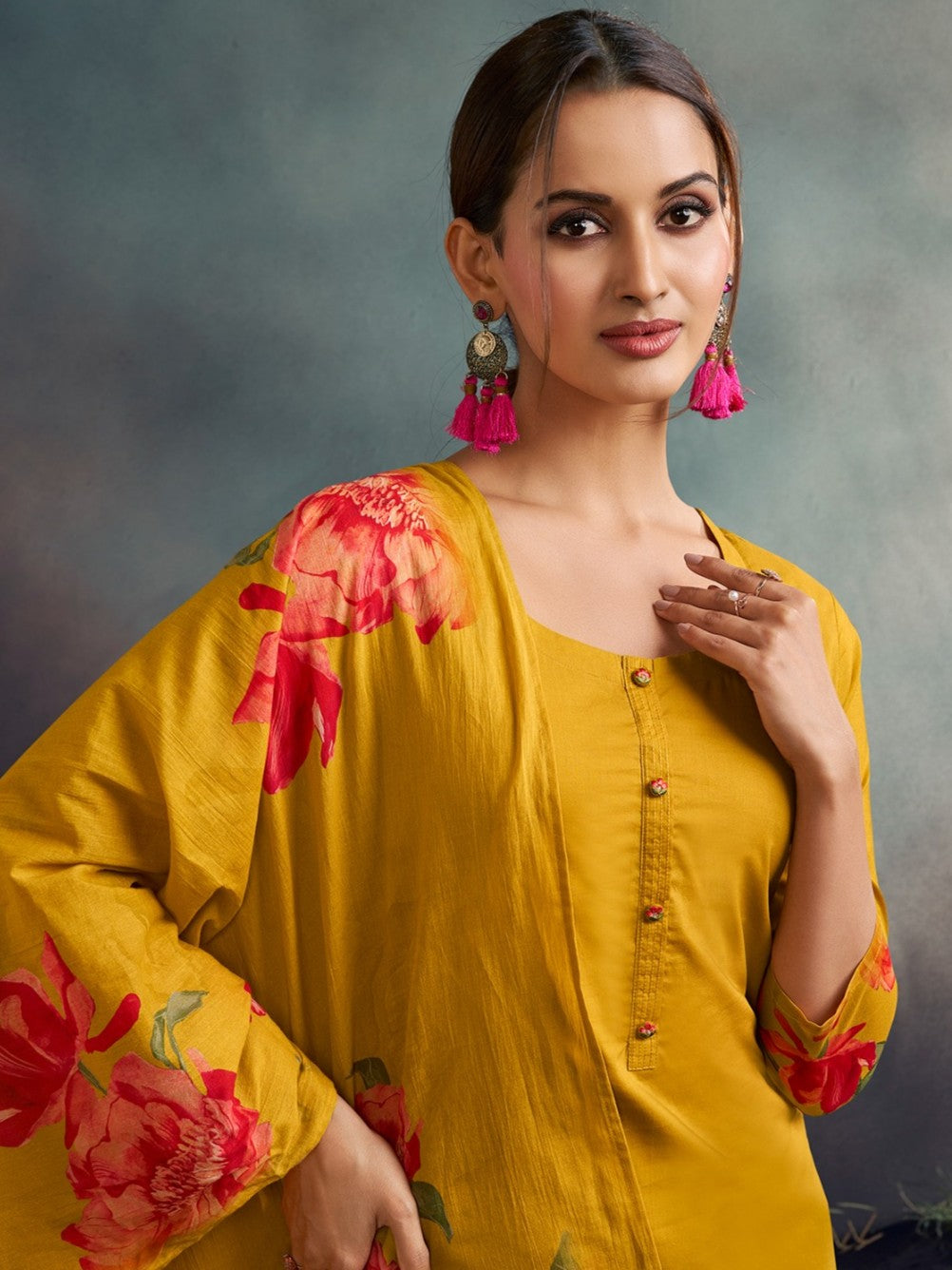 Woman wearing a yellow floral outfit with pink earrings against a gray background